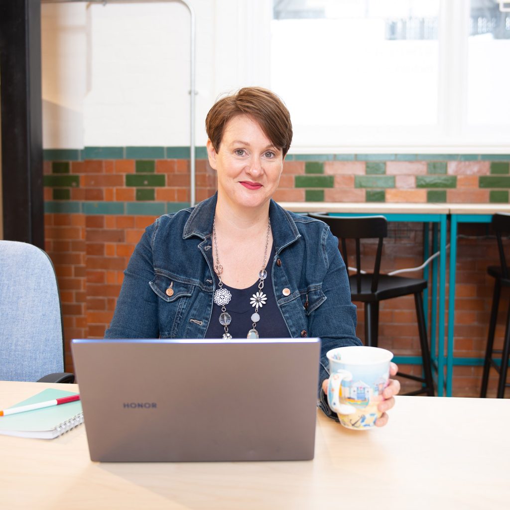 Catherine Jones wearing denim jacketfacing camera sat at desk with laptop and cup of tea